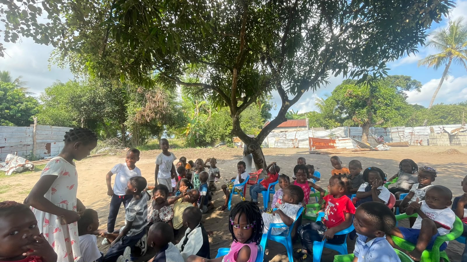 Crianças reunidas em cadeiras coloridas sob a sombra de uma árvore